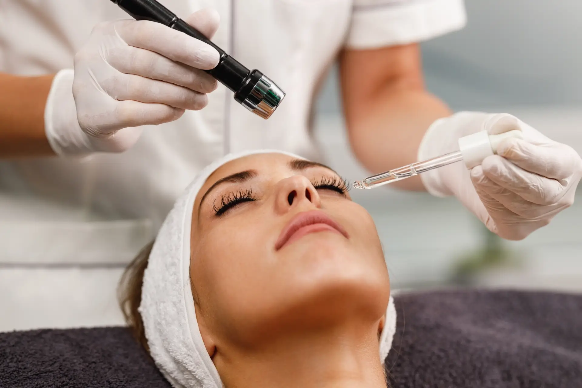 Gloved hands hold a pen-style treatment handpiece above the face of a client resting with a white headband and closed eyes
